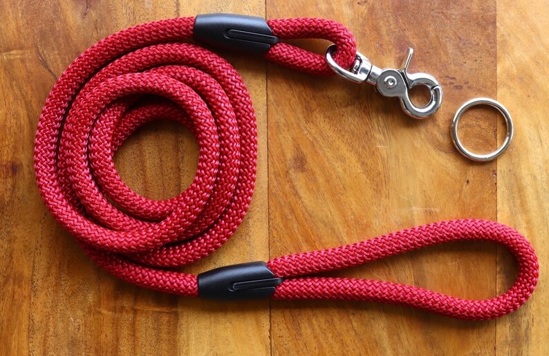 May include: Red rope dog leash with a black handle and a silver clip. The leash is coiled up on a wooden surface.