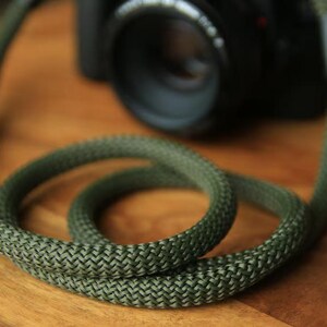 Peut inclure: Gros plan d'une sangle d'appareil photo verte avec une texture tissée, enroulée sur une surface en bois. Un appareil photo noir est partiellement visible en arrière-plan. La sangle suggère une bonne résistance.