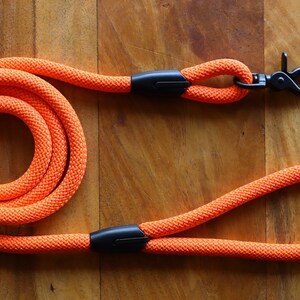 May include: Bright orange dog leash made of thick, woven rope. The leash features a black metal clasp and a loop handle. The rope is coiled in a circular pattern. The leash is designed for pet owners.