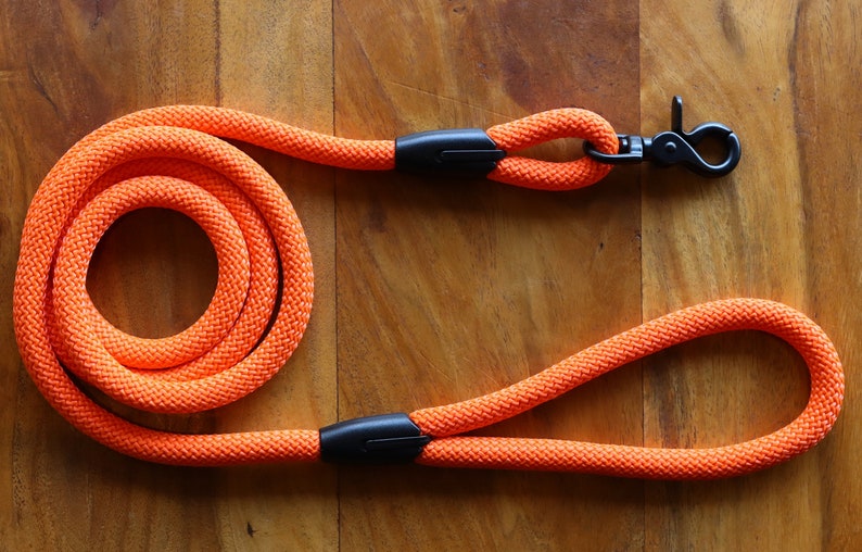 May include: Bright orange dog leash made of thick, woven rope. The leash features a black metal clasp and a loop handle. The rope is coiled in a circular pattern. The leash is designed for pet owners.