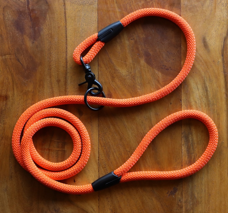 May include: Bright orange dog leash with a black metal clasp and handle. The leash is made of a thick, woven rope material. The handle and clasp are connected by a black plastic piece. The leash is coiled on a wooden surface.