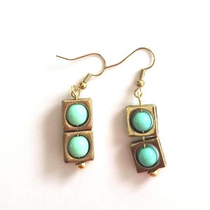 Puede incluir: Un par de pendientes colgantes con dos marcos cuadrados dorados apilados, cada uno con una cuenta redonda y lisa de color turquesa claro. Los pendientes cuelgan de ganchos de metal dorado e incluyen una pequeña cuenta dorada en la base.