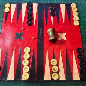 May include: A red and black backgammon board with wooden game pieces and dice. The board features a central black stripe with the text "ATELIER OZVOZ". The game pieces are black and cream-colored.