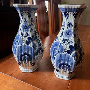 May include: A pair of white ceramic vases with a hexagonal shape and a fluted neck. Each vase features a hand-painted blue floral pattern with intricate petals, leaves, and scrolling details covering the exterior surface from base to rim.