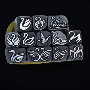 May include: A collection of small, dark gray square stamps, each featuring a white swan design. The stamps are arranged in a grid and held together with a yellow rubber band. The background is solid black.