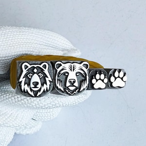 Puede incluir: Un juego de tres sellos de goma con diseños de osos. Los sellos incluyen dos caras de oso y dos huellas de patas de oso. Los sellos son en blanco y negro y están montados sobre una base de madera. Los sellos son sostenidos por una mano enguantada.