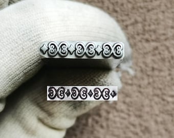 May include: A silver-toned metal stamping tool featuring a repeating decorative pattern of three stylized scrollwork motifs separated by small diamond shapes. The tool has a rectangular face that leaves a clear, embossed impression of the design.