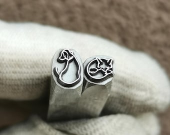 May include: Two silver-colored metal stamping tools with square shafts. One tool features a raised outline of a sitting dog, while the other tool features a raised outline of a cat head with pointed ears, both designed for creating impressions.