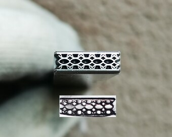 May include: A silver-toned metal stamping tool featuring a rectangular head with a repeating geometric pattern of small, interconnected oval shapes. Below the tool head, a separate rectangular sample shows the embossed impression of the design.