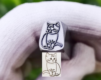 May include: A silver-toned metal stamping tool featuring a recessed cat design on its square face. The tool is shown alongside a clear black and white ink impression of the same cat illustration, which depicts a seated feline with distinct facial markings.
