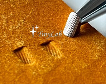 May include: A silver metal leather stamping tool with a textured, diamond-patterned tip is pressing into a piece of tan leather, creating a small, grooved impression. The text TroyLab is printed in white near the tool tip on the leather surface.