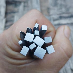 Könnte beinhalten: Eine Sammlung quadratischer Stempel aus Metall, die in einer Hand gehalten werden. Die Stempel sind silberfarben und haben verschiedene Größen.