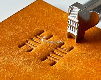 May include: A silver metal leathercraft stamping tool with a textured tip is pressed into a tan leather surface, creating two parallel rows of embossed, ribbed impressions. The tool features a cylindrical handle and the brand name TroyLab is visible.