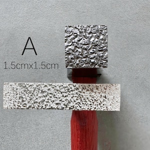 Może przedstawiać: Srebrna metalowa pieczątka o teksturowanej powierzchni, o wymiarach 1,5 cm x 1,5 cm. Pieczątka jest przymocowana do czerwonego drewnianego uchwytu. Litera "A" jest wydrukowana nad pieczątką.