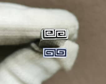 May include: A metal stamp tool featuring a rectangular head with a raised geometric Greek key pattern. The stamp is shown alongside a small, rectangular white impression block that displays the same dark blue Greek key design clearly.