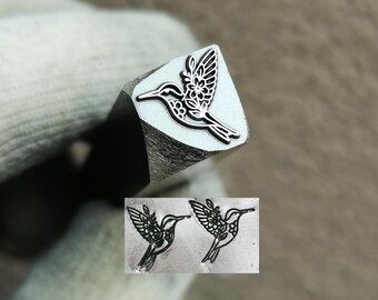 May include: A silver-colored metal stamping tool featuring a hummingbird design with floral patterns on its head. The end of the tool shows the raised hummingbird shape, and an inset image displays two clear impressions of the hummingbird stamp.