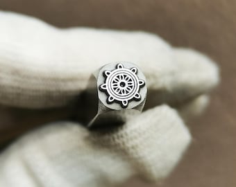 May include: A silver-toned metal stamping tool featuring a circular head with an engraved ship's wheel design. The stamp has a square-shaped shaft and a detailed, symmetrical pattern consisting of a central flower-like motif surrounded by spokes.