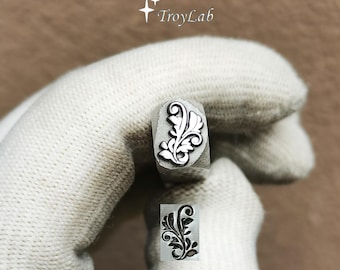 May include: A metal stamping tool featuring a detailed floral scroll design on its tip, with the TroyLab logo visible above. The stamp head displays a raised, intricate leaf pattern, and a clear black ink impression of the same design is shown below.