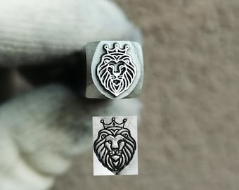 May include: A metal stamping tool featuring a square head with a recessed lion head design wearing a crown. Below the tool, a clear black and white impression of the same lion head with a crown is visible, showing the detailed engraved lines.