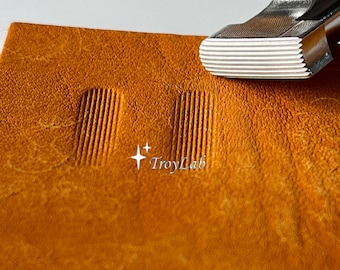 May include: A metal leather stamping tool with a rectangular, grooved head positioned above a piece of tan leather. The leather displays two identical, parallel rectangular impressions with fine vertical lines, and the text TroyLab is visible.
