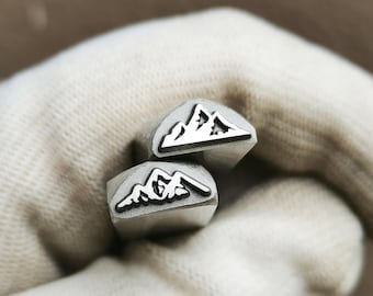 May include: Two silver-colored metal stamping tools, each featuring a raised mountain range design on the flat end. The tools have a square-shaped shaft and a metallic finish, designed for creating impressions in materials through manual pressure.