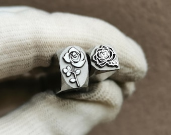 May include: Two silver-toned metal stamping tools with square heads. One tool features an engraved design of a rose on a stem with leaves, while the other tool displays a detailed, intricate rose blossom design with visible petals and foliage.