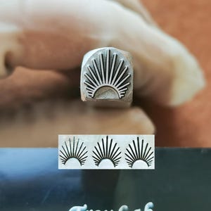 May include: A metal stamp featuring a sunburst design, held by a gloved hand. The stamp's impression is visible below, alongside the text "TroyLab". The sunburst design consists of a semi-circle with radiating lines.