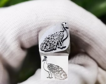 May include: A silver-colored metal stamping tool featuring a detailed engraved peacock design on its flat end. The tool is shown with a clear black ink impression of the peacock motif directly below the metal head, displaying the bird's plumage.