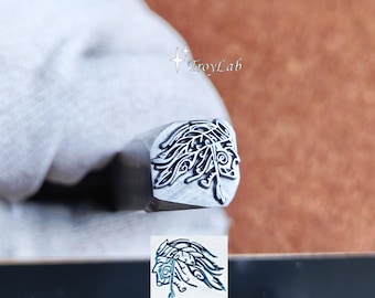 May include: A metal stamping tool featuring a detailed engraving of a profile of a person wearing a feathered headdress on its square face. The text TroyLab is visible near the stamp and on a black surface below with a printed logo.