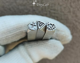 May include: Three silver-colored metal stamping tools with square shafts, each featuring a distinct engraved design on the end: an anchor, a triangle with a smaller triangle inside, and an eye with radiating lines. The text TroyLab is visible.