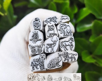 May include: A collection of twelve metal stamps featuring engraved animal designs including an eagle, wolf, dolphin, cat, elephant, butterfly, deer, horse, tiger, peacock, and lion. The stamps are arranged in a grid and shown with a printed reference sheet.