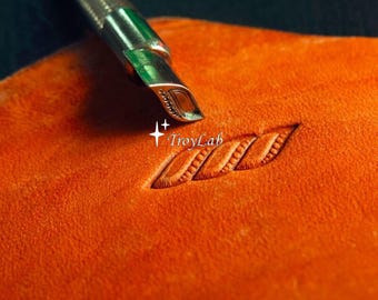 May include: A metal leather stamping tool with a textured handle and a curved tip is pressing into a piece of orange leather, creating a repeating rope-like pattern. The text TroyLab is visible near the stamped design on the leather surface.