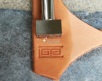 May include: A silver metal leather stamping tool with a rectangular head featuring a Greek key pattern. The tool is positioned on a piece of tan leather, showing a clear, debossed impression of the geometric design. The text TroyLah is visible.