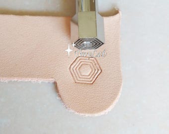 May include: A silver-colored metal leather stamping tool with a hexagonal tip, positioned above a piece of light tan leather that features a stamped hexagonal pattern and the text TroyLab printed in white.