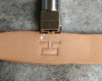 May include: A silver metal leather stamping tool pressing a T-shaped design into a strip of light tan leather. The stamp features the text TroyLab with two small star icons above the letter T, creating a clear, debossed impression in the material.