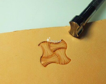 May include: A metal leather stamping tool with a textured cylindrical handle positioned next to a tan leather surface. The leather features a stamped impression of a four-part woven pattern with curved edges and fine parallel lines within each section.