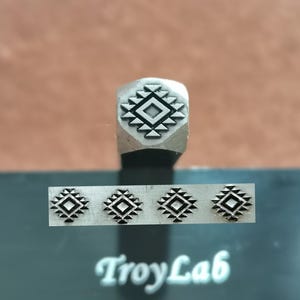 Könnte beinhalten: Ein Metallstempel mit einem geometrischen Design, das ein zentrales Quadrat umgeben von Dreiecken aufweist. Darunter sind vier gestempelte Abdrücke desselben Designs zu sehen. Der Text "TroyLab" ist weiß auf einer dunklen Oberfläche gedruckt.
