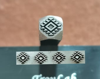 May include: A metal stamp with a geometric design, featuring a central square surrounded by triangles. Below, four stamped impressions of the same design are visible. The text "TroyLab" is printed in white on a dark surface.