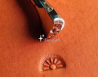 May include: A silver-colored metal leather stamping tool with a semi-circular head featuring a sunburst pattern. The tool is positioned above a matching semi-circular sunburst impression stamped into orange leather. The text TroyLab is visible.