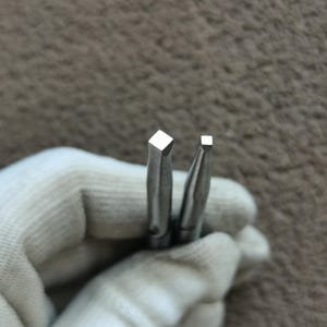 May include: Two silver-colored metal tools with square-shaped tips. The tool on the left features a larger square tip, while the tool on the right has a smaller square tip. Both tools have tapered shafts and a smooth, metallic finish.