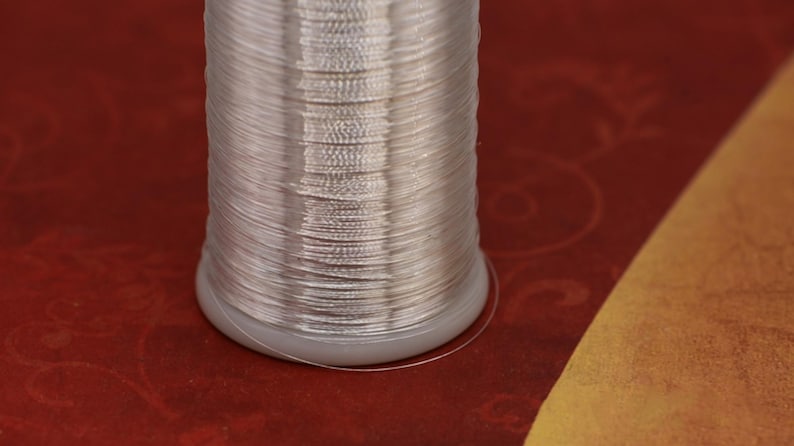 Może przedstawiać: Szpula srebrnej nici metalicznej, ciasno nawinięta na białą plastikową szpulkę.