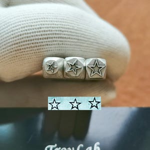 May include: Three metal star stamps of varying sizes, each with a five-pointed star design. The stamps are held by a gloved hand. Below the stamps is a line of three star imprints and the text "TroyLab".