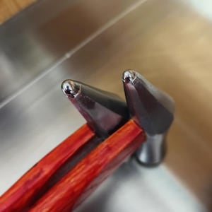 Puede incluir: Dos cabezales de martillo de metal con mangos de madera roja. Los cabezales de martillo son puntiagudos y tienen un acabado brillante.