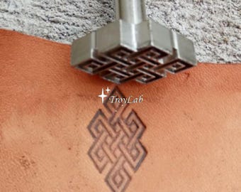 May include: A silver-colored metal leather stamping tool with a geometric knot design. The tool is positioned above a tan leather surface, which displays a clear, dark brown embossed impression of the same intricate knot pattern and the text TroyLab.