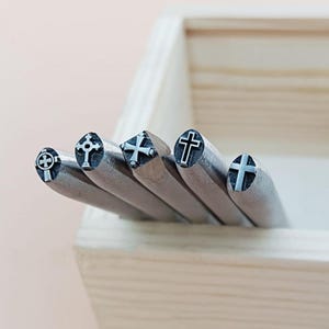 Op de afbeelding: Vijf metalen stempelgereedschappen met verschillende kruisontwerpen in een houten doos. De gereedschappen worden gebruikt om metaal te stempelen.