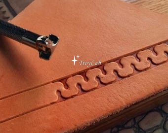 May include: A metal leather stamping tool with a textured handle positioned above a piece of tan vegetable-tanned leather. The leather features a continuous, embossed wavy line pattern created by the tool. The text TroyLab is visible.