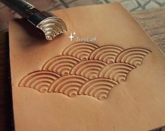 May include: A metal leather stamping tool with a semi-circular head featuring concentric arc patterns. The tool is positioned on a piece of light tan leather that displays a repeating, embossed wave-like pattern. The text TroyLab is visible.