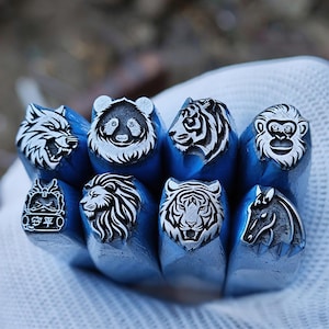 Könnte beinhalten: Eine Sammlung von Metallstempeln mit eingravierten Tiergesichtern. Die Stempel zeigen einen Wolf, Panda, Tiger, Affen, Löwen, Tiger und ein Pferd. Die Stempel werden in einer behandschuhten Hand gehalten.