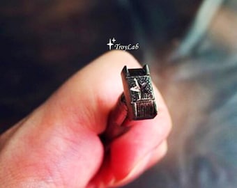 May include: A small, dark metallic coil component featuring a textured surface with a central star emblem. The piece is marked with the text TroyLab and displays a series of fine, tightly wound wire wraps along its rectangular structure.