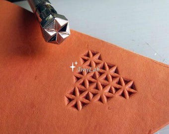 May include: A silver metal leather stamping tool with a hexagonal head featuring a star pattern, positioned above a piece of tan leather that displays a triangular arrangement of stamped star impressions and the text TroyLab in white.